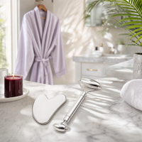 Silver facial roller and gua sha on marble countertop with robe and candle